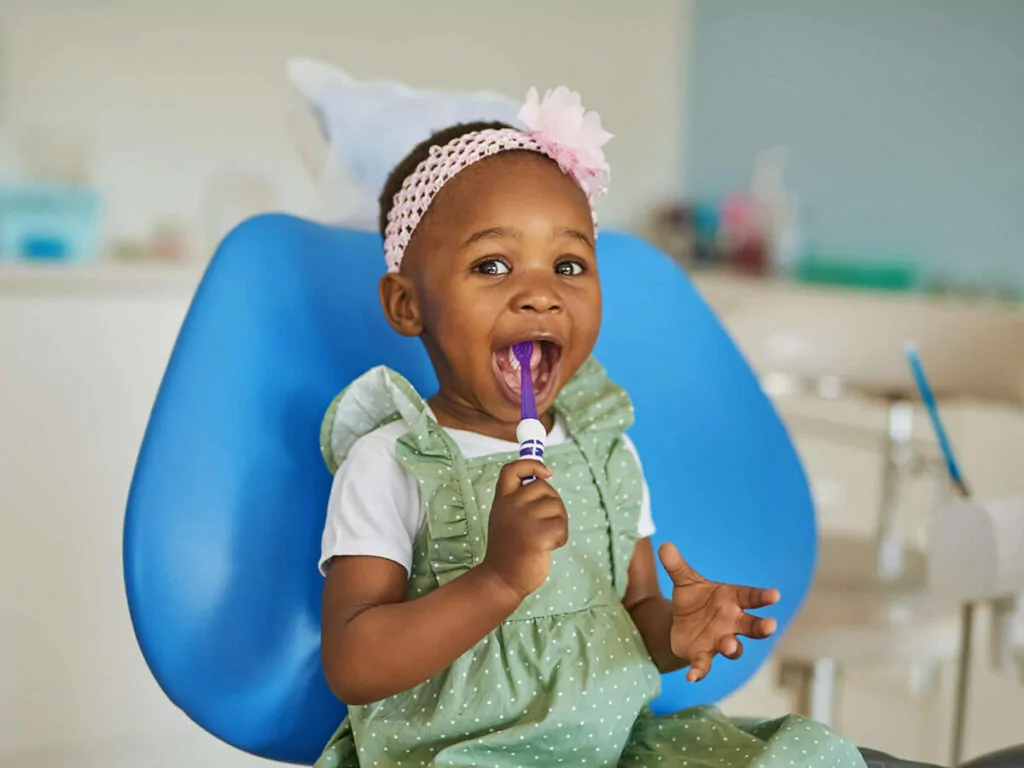 A young girl sits in a dental exam chair with a toothbrush in her mouth at a pediatric dentistry appointment